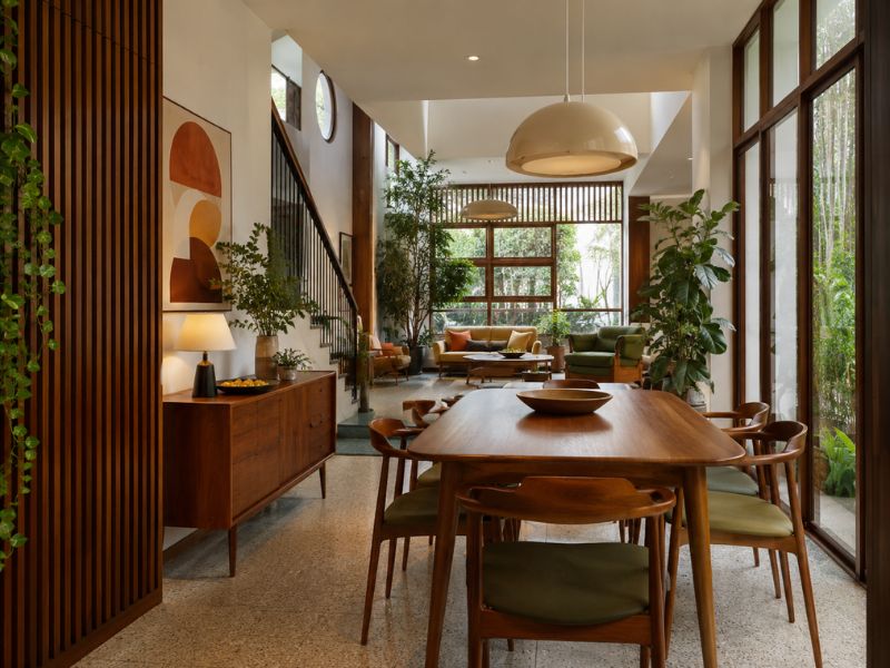 Khu bàn ăn liền phòng khách phong cách thiết kế nội thất Mid-Century Modern với bàn ăn gỗ dài, ghế chân thanh, đèn thả mái vòm và nhiều cây xanh