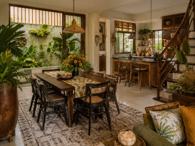 Khu bàn ăn và bếp phong cách Tropical Maximalist với bàn gỗ tối màu, ghế mây, hoa văn vải và nhiều cây xanh