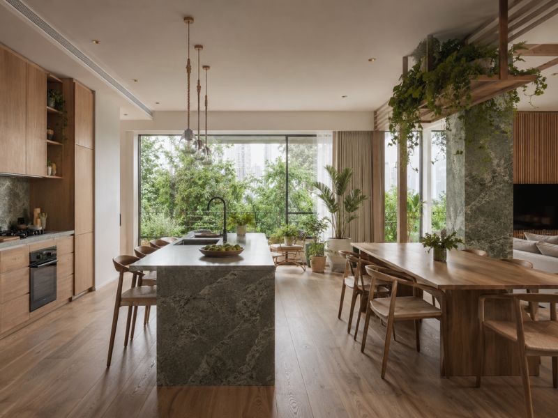 Khu bếp và bàn ăn trong các phong cách thiết kế nội thất chung cư Biophilic với đảo bếp đá, bàn ăn gỗ dài, cửa mở lớn và nhiều cây xanh xung quanh