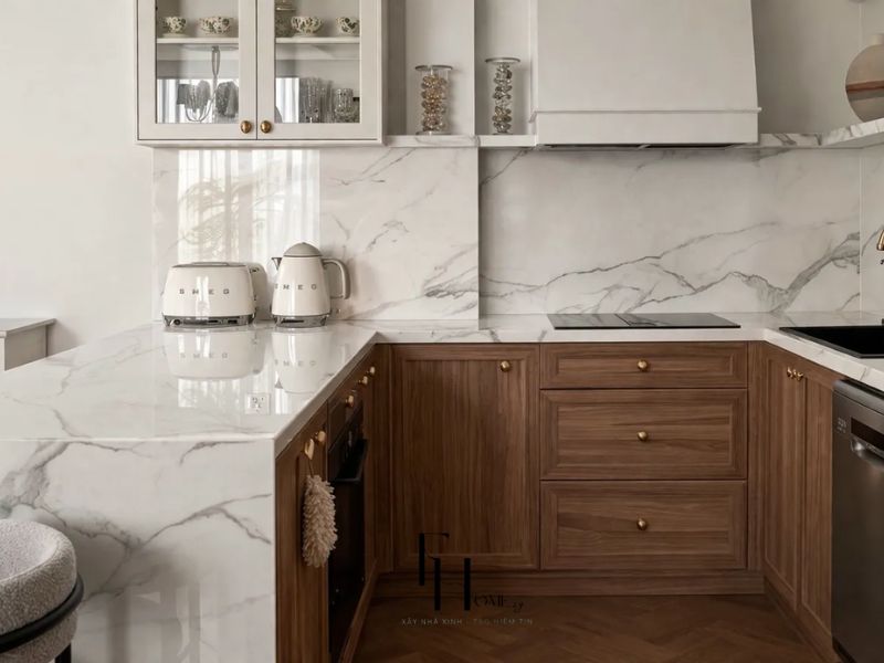 Phòng bếp chung cư 55m2 phong cách Scandinavian với tủ gỗ walnut nâu tay nắm vàng gold, mặt bếp đá marble trắng và thiết bị SMEG kem
