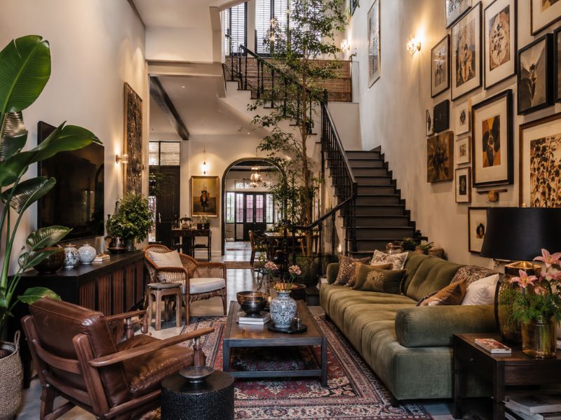 Phòng khách nhà phố phong cách Eclectic với sofa xanh rêu dài, ghế da nâu, tường treo tranh gallery, cầu thang sắt và cây xanh trang trí