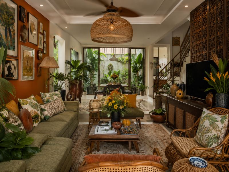 Phòng khách phong cách Tropical Maximalist với sofa xanh olive, tường nhấn cam đất, nhiều tranh treo và cây xanh nhiệt đới