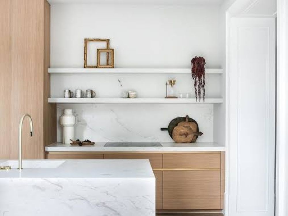 Bếp tối giản với đảo bếp đá trắng, tủ dưới gỗ sáng và kệ ngang treo tường