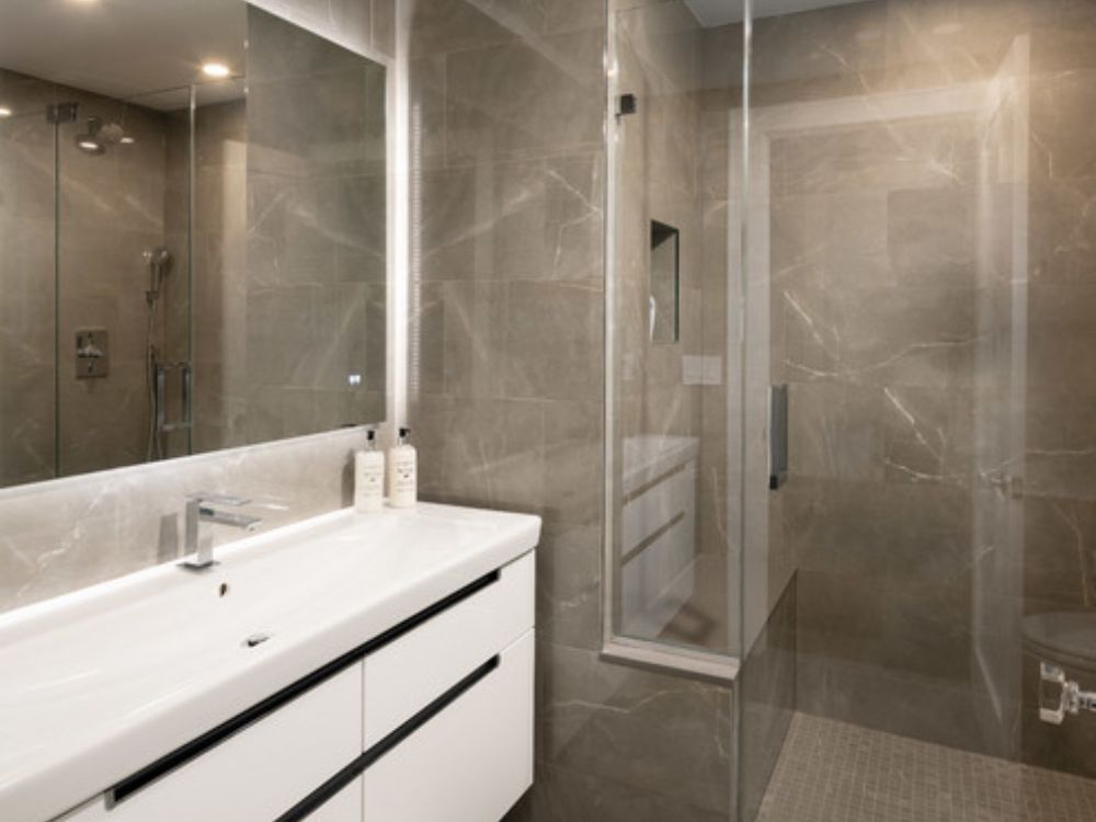 Nhà tắm hiện đại với bồn rửa trắng dài, tường ốp đá marble xám bóng, buồng tắm kính