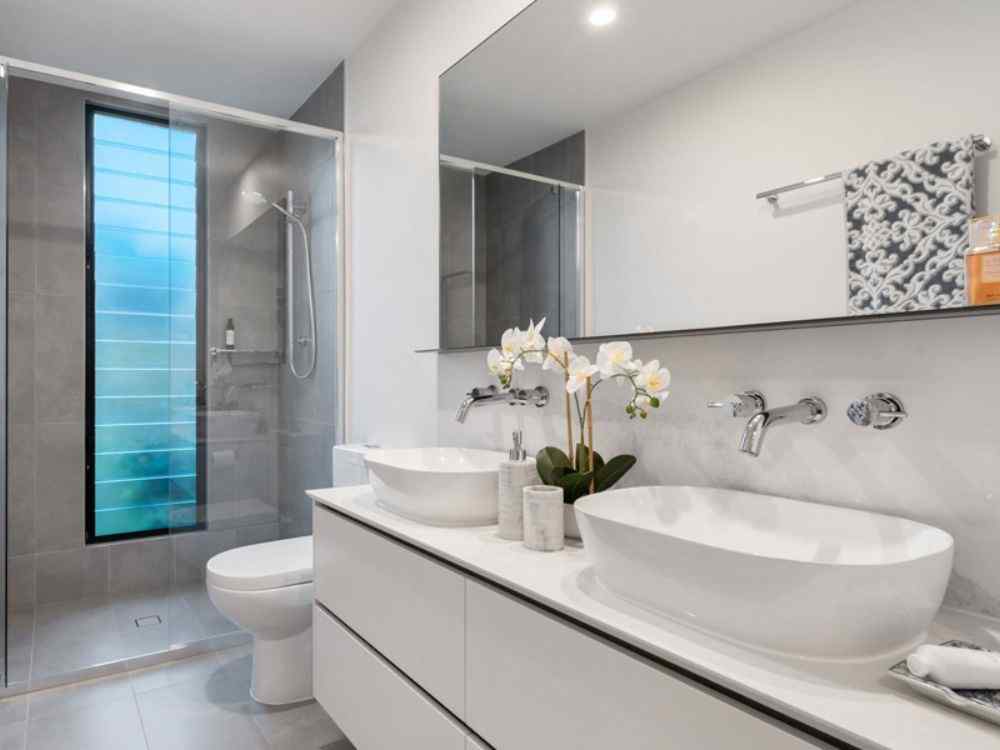 Nhà vệ sinh hiện đại tông xám trắng với bồn rửa đôi, gương lớn và ô cửa kính dọc đón sáng tự nhiên