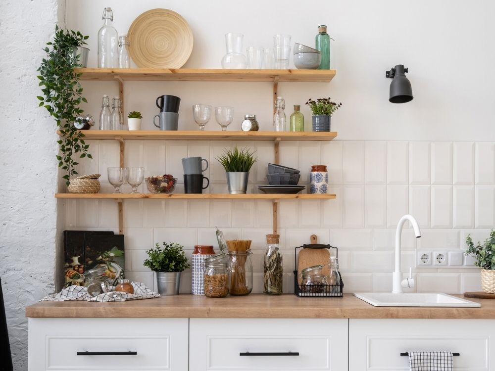 Phòng bếp trắng với kệ gỗ mở, vật dụng thủy tinh và cây xanh tạo cảm giác ấm cúng