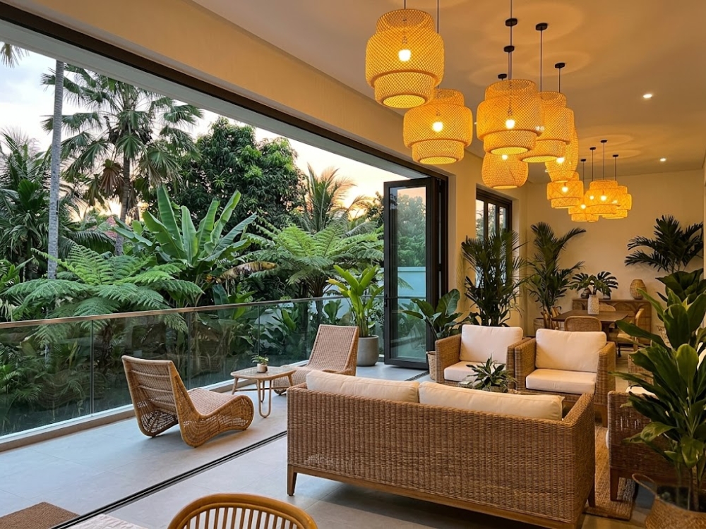 Phòng khách hoặc ban công Tropical Modern — mây, cây nhiệt đới lớn, đèn mây đan, ánh sáng vàng ấm.