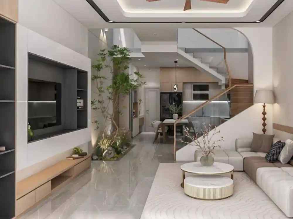 Phòng khách liền bếp thoáng mở, sofa L be nhạt, bàn đá tròn, cầu thang kính, tiểu cảnh xanh nổi bật