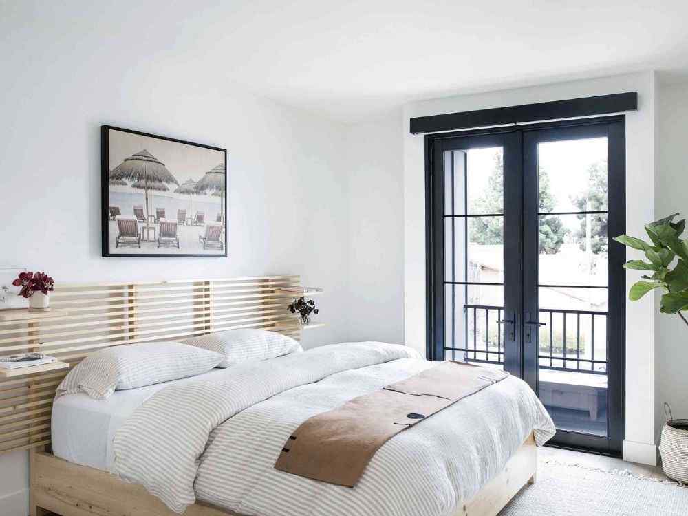 Phòng ngủ sáng sủa với giường gỗ sáng, cửa kính đen lớn mở ra ban công và cây xanh trang trí