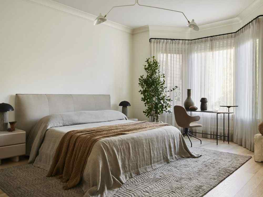 Phòng ngủ tối giản với giường lớn phủ chăn nâu, rèm trắng mềm rũ và cây xanh góc cửa sổ
