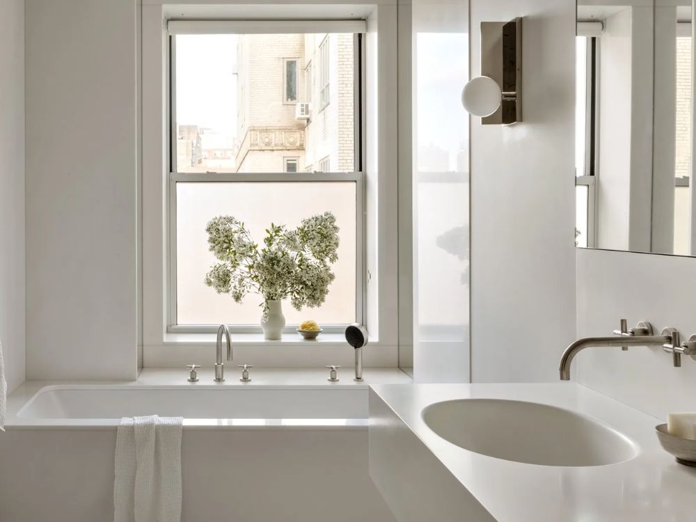 Phòng tắm tối giản với bồn tắm vuông trắng, cửa sổ lớn và lọ hoa nhỏ trang trí nhẹ nhàng