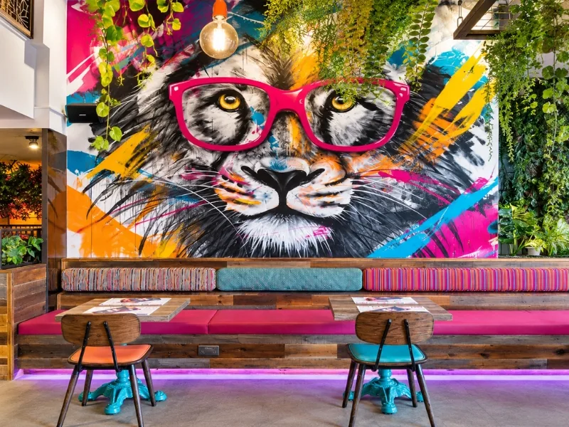 Quán cà phê phong cách Funky có tranh mèo graffiti khổ lớn phía sau ghế băng hồng và bàn nhiều màu.