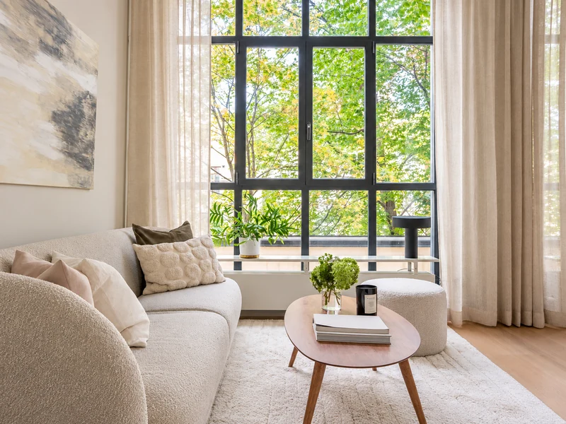 Phòng khách chung cư có cửa sổ kính lớn, sofa be, bàn trà tròn và rèm voan
